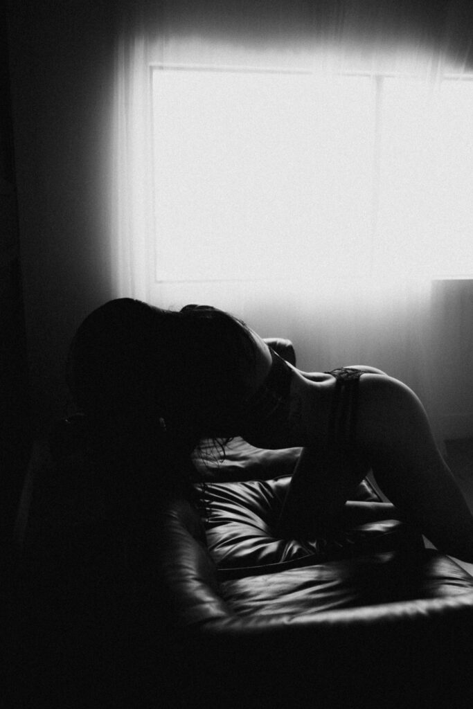 Black and white boudoir photograph of a woman posing on a leather couch in soft window light inside a boudoir photography studio.