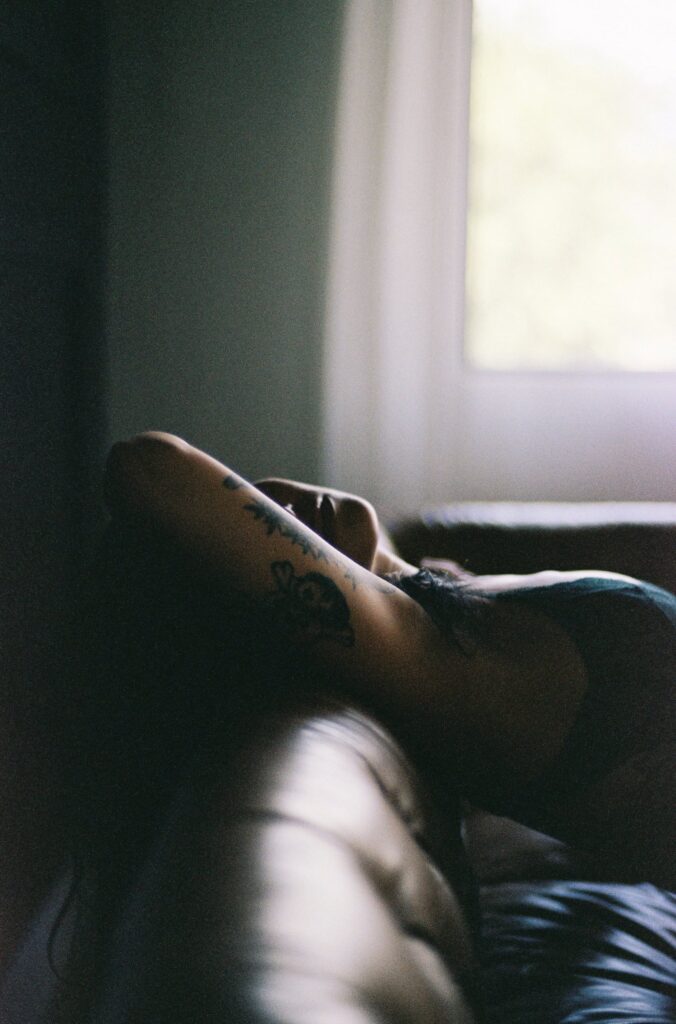 Soft, intimate boudoir portrait of a woman reclining on a leather couch near window light inside a Phoenix boudoir photography studio.